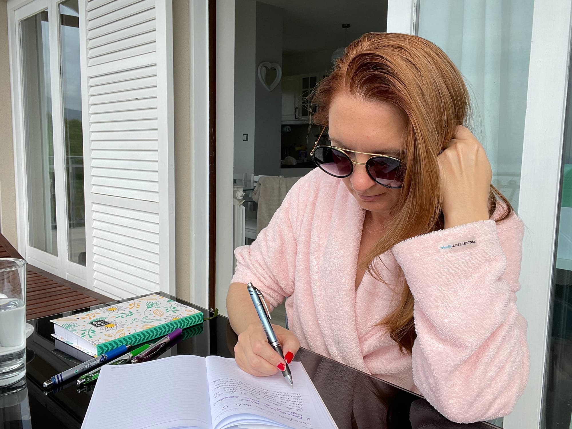 Frau mit langen blond-roten Haaren, Sonnenbrille im rosa Bademantel sitzt am Tisch und schreibt im Notizbuch.